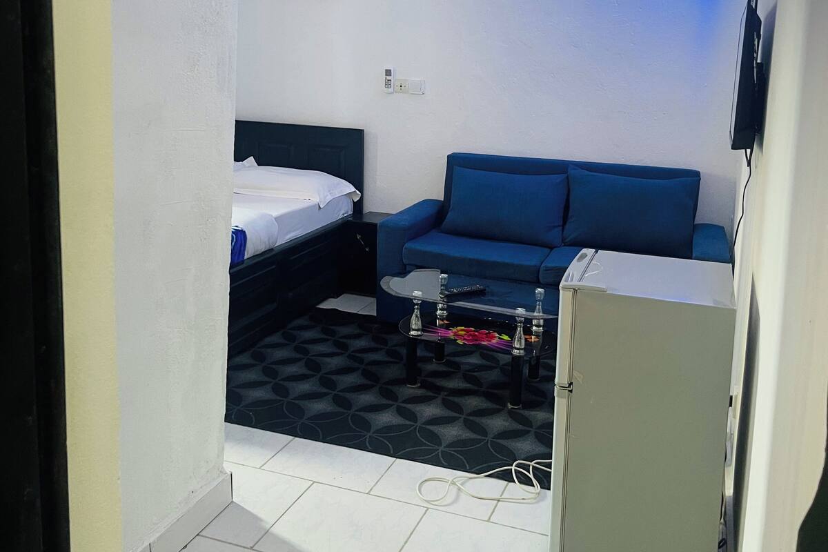 A compact hotel room with a bed, a blue sofa, a small table, and a refrigerator.