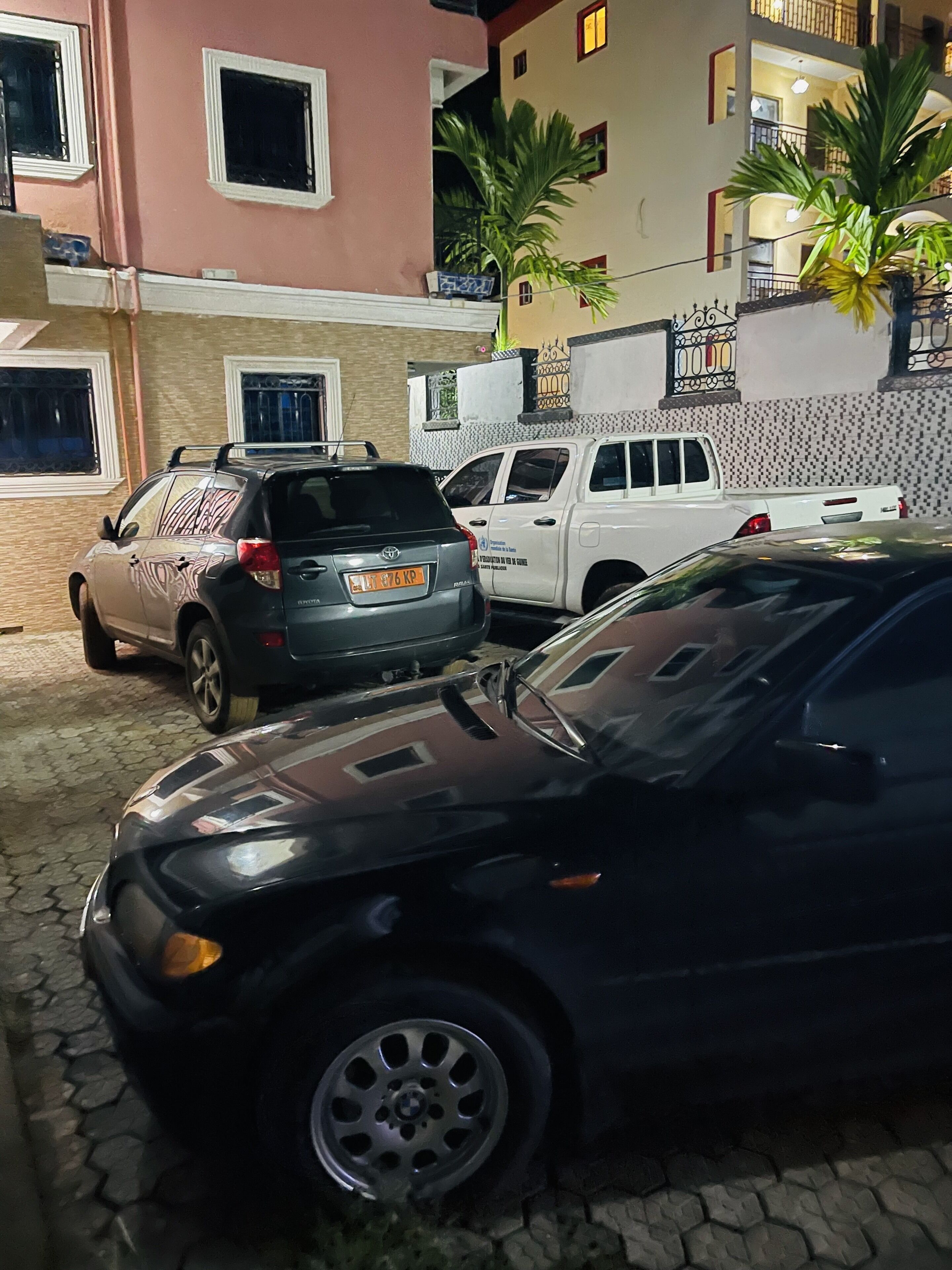Un estacionamiento con varios coches, incluyendo un SUV negro y una camioneta blanca, frente a un edificio con fachada rosa y un balcón.