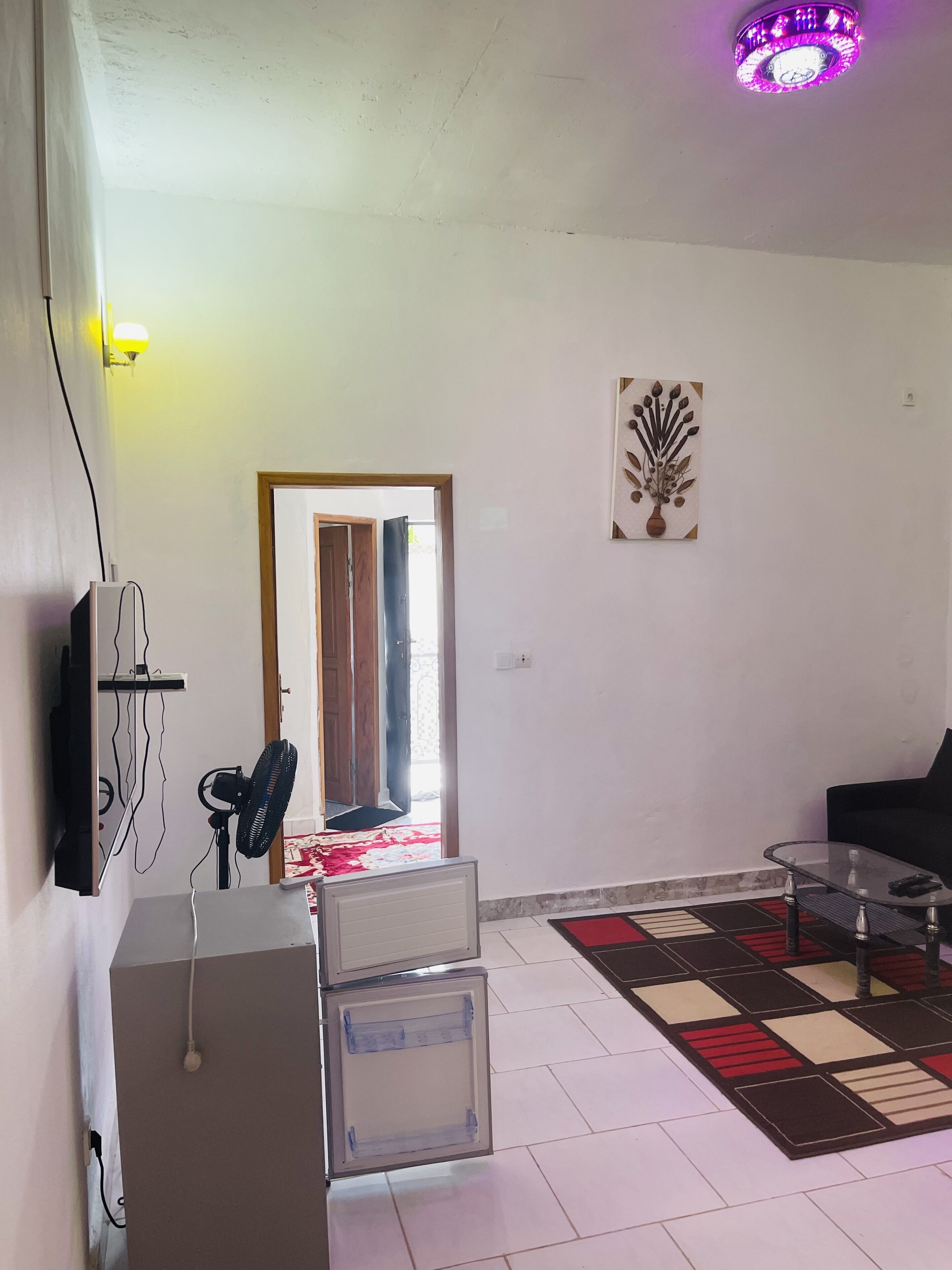 Una sala de estar con un radiador, un ventilador, una mesa de centro de vidrio, un sofá negro, un tapete estampado y una puerta que da al exterior.