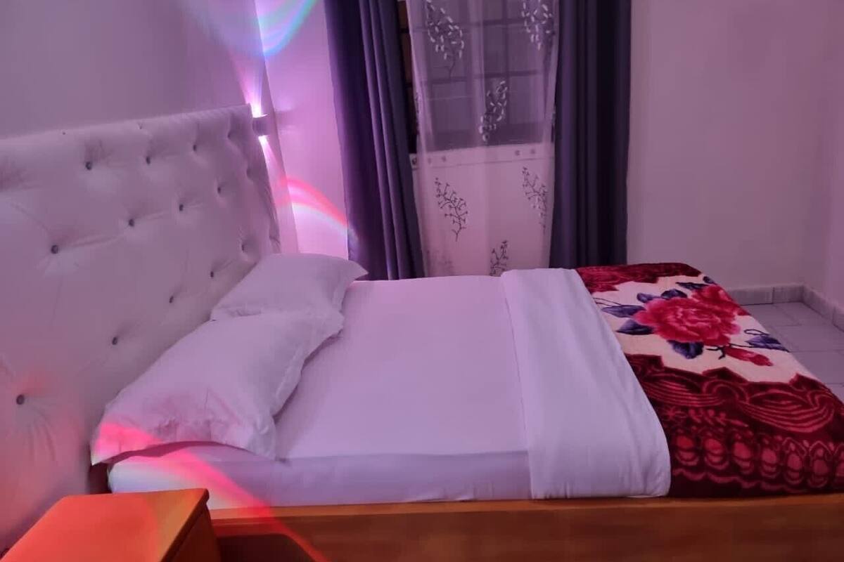 A bedroom with a tufted headboard, a bed with white and red floral bedding, a wooden nightstand, and a window with sheer curtains.