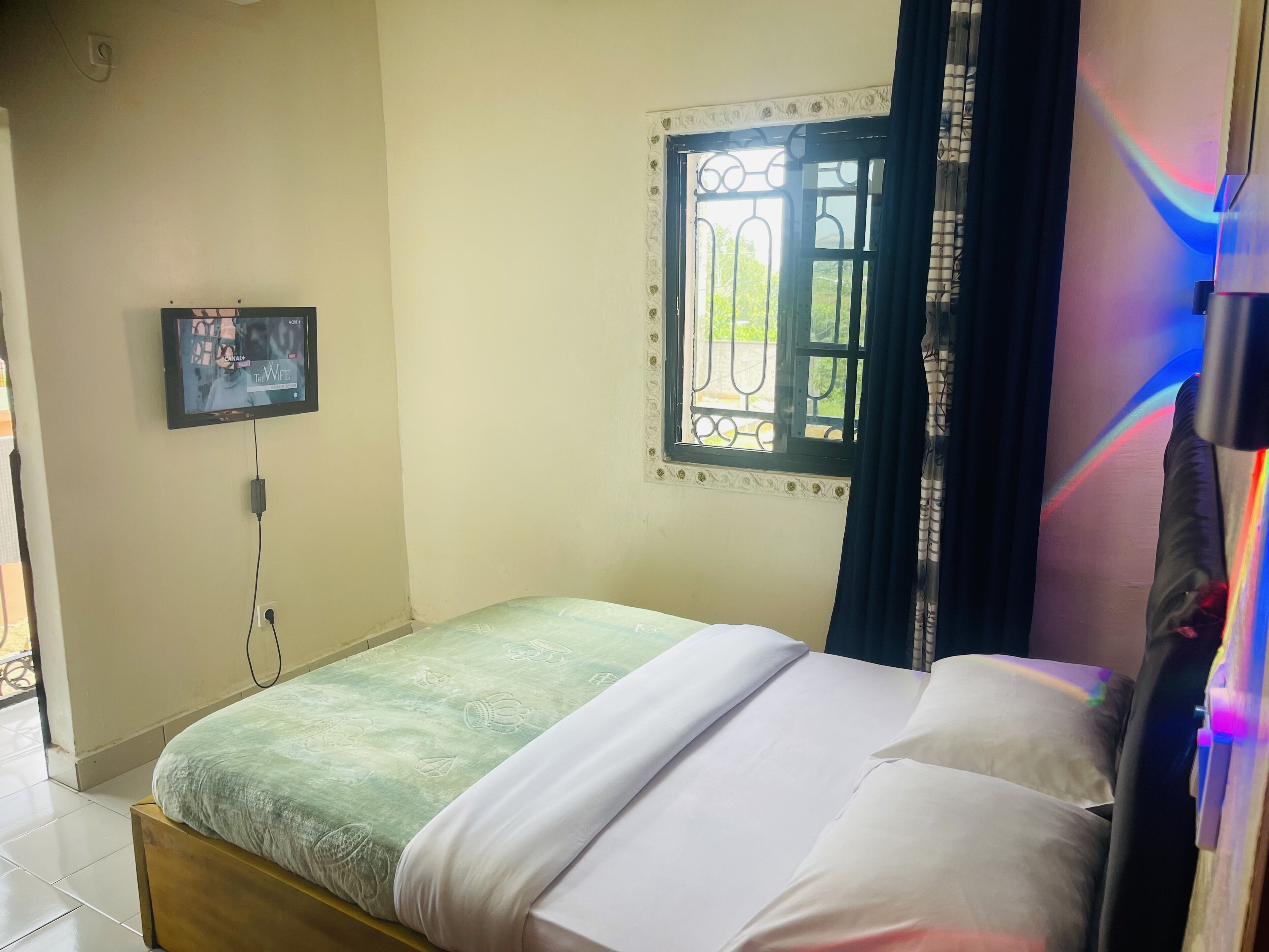 A bedroom with a bed, a TV mounted on the wall, a window with decorative iron bars, and a curtain.