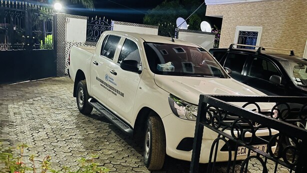 A white pickup truck parked in a driveway with a tiled patio area, a metal gate, and a building with a pink facade in the background. - Hotel Svet Residence (Kribi)