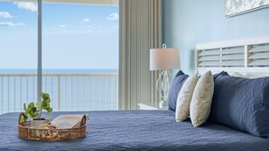 A bedroom with a view of the ocean, a bed with blue bedding, a bedside table with a lamp, and a wicker basket with a plant. - Stunning Oceanfront Condo! Sleeps 8, Modern Appliances, Majestic (Panama City Beach)