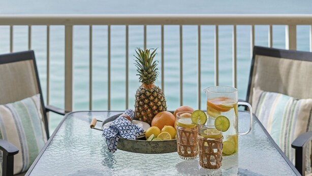 A table with a glass pitcher, glasses, a pineapple, oranges, and a bowl of fruit on a balcony with a view of the sea. - Stunning Oceanfront Condo! Sleeps 8, Modern Appliances, Majestic (Panama City Beach)