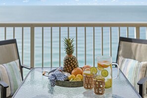 A table with a glass pitcher, glasses, a pineapple, oranges, and a bowl of fruit on a balcony with a view of the sea.