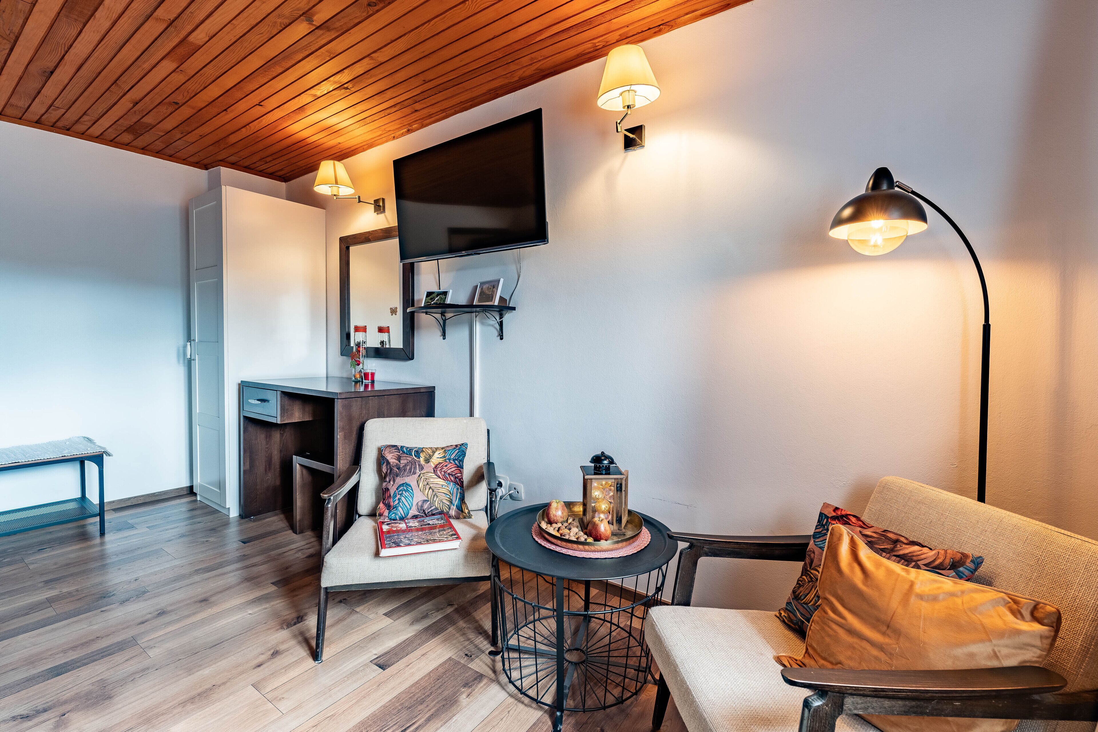 Ein modernes Wohnzimmer mit Holzboden, einem Wand-Fernseher, einem runden Couchtisch mit Dekoration, einer Couch mit Kissen und einem Schreibtisch mit Stuhl.