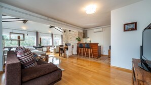 Ein modernes Wohnzimmer mit einem braunen Sofa, Holzboden, einer Essecke mit Stühlen, einer Küche mit Steinwand und einem Fernseher auf einem Holzbodenständer.