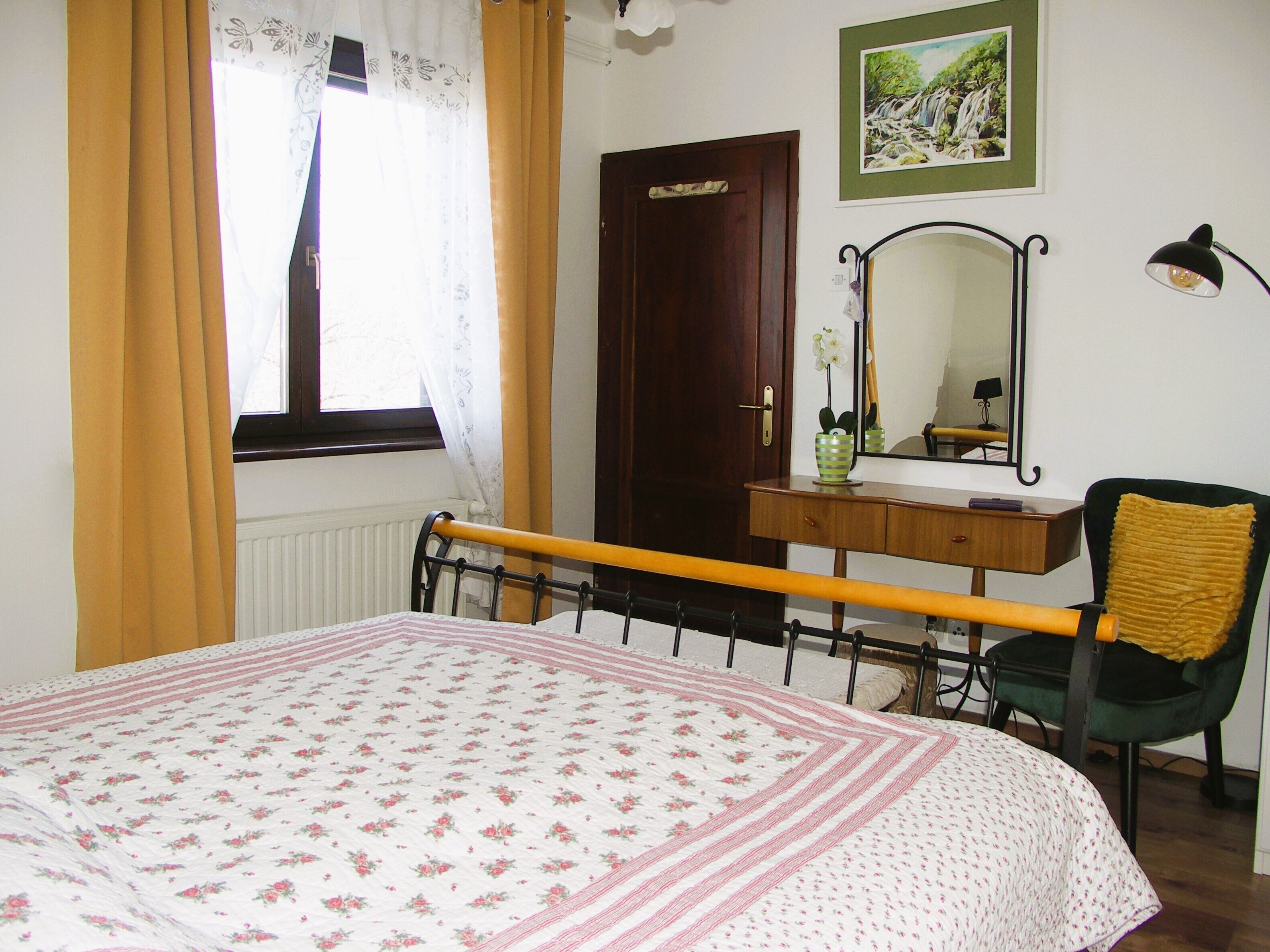 A bedroom with a bed, a dresser, a chair, a window with curtains, a door, and a mirror on the wall.