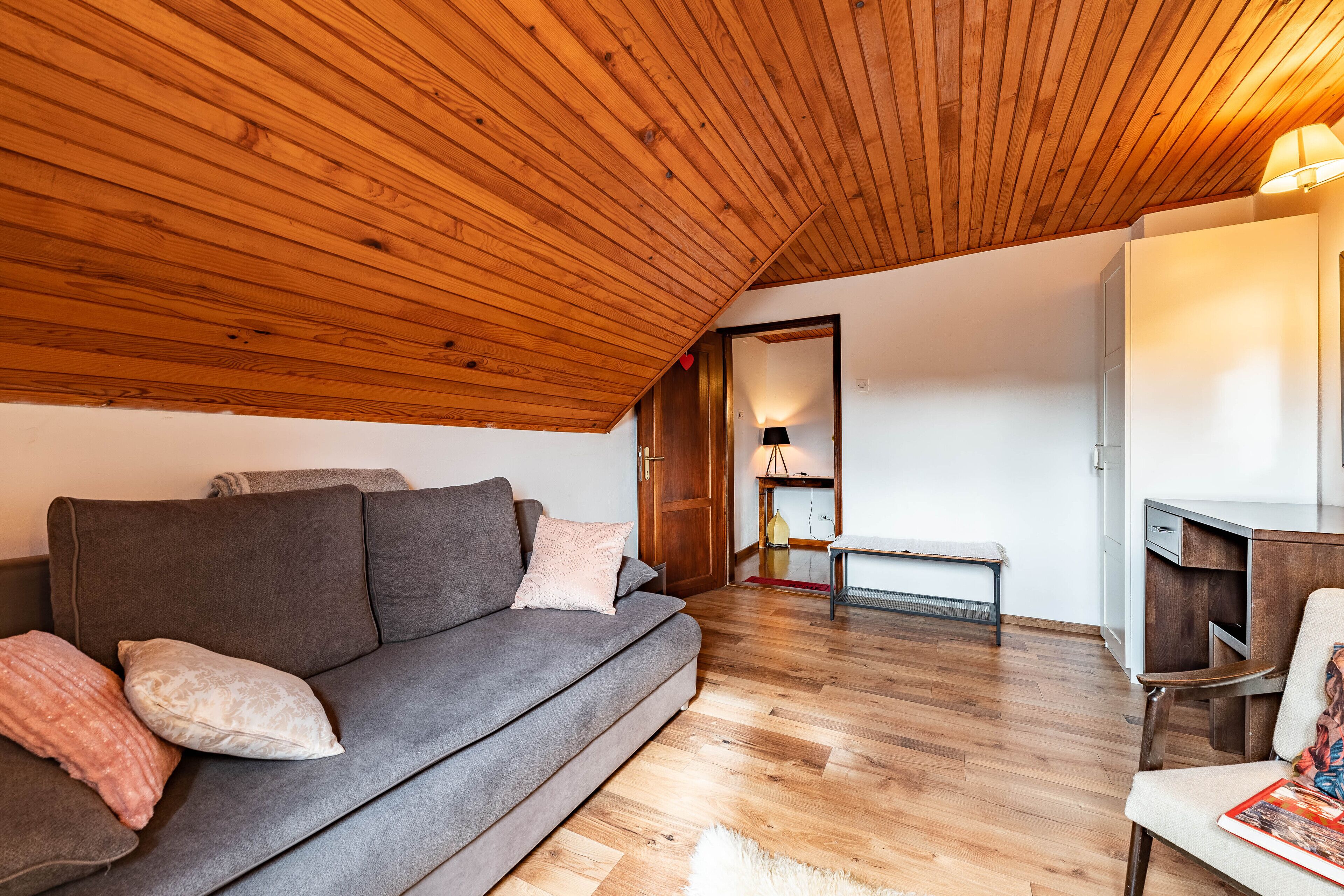 Un salón acogedor con techo de madera, un sofá gris con cojines, suelo de madera y una mesita con una lámpara.