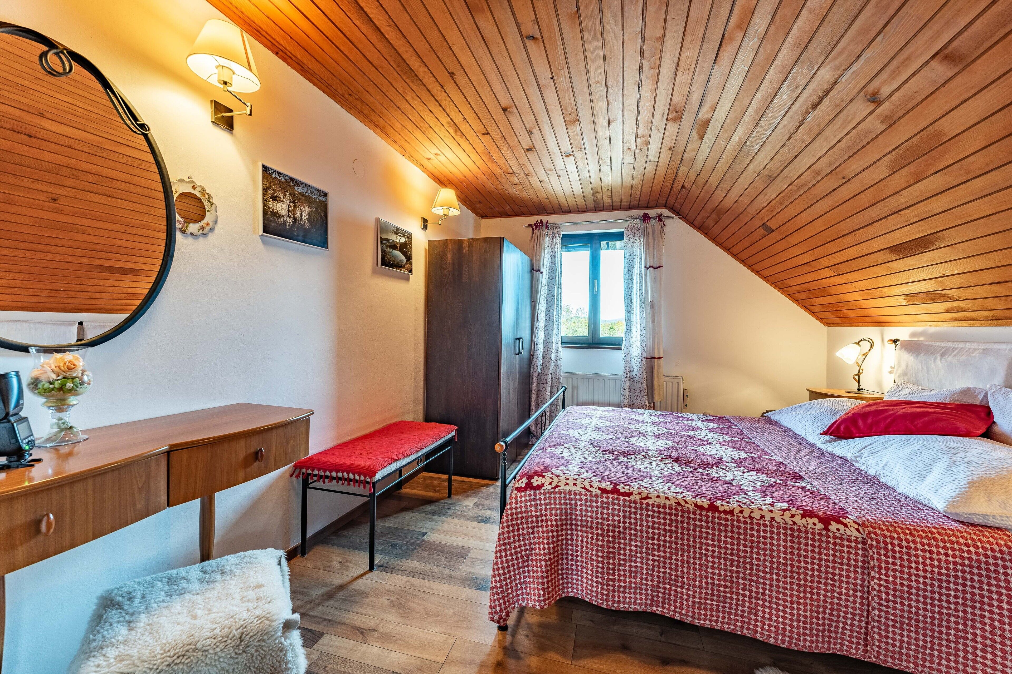 A bedroom with a wooden ceiling, a bed with a red and white checkered bedspread, a bedside table with a lamp, and a bench.