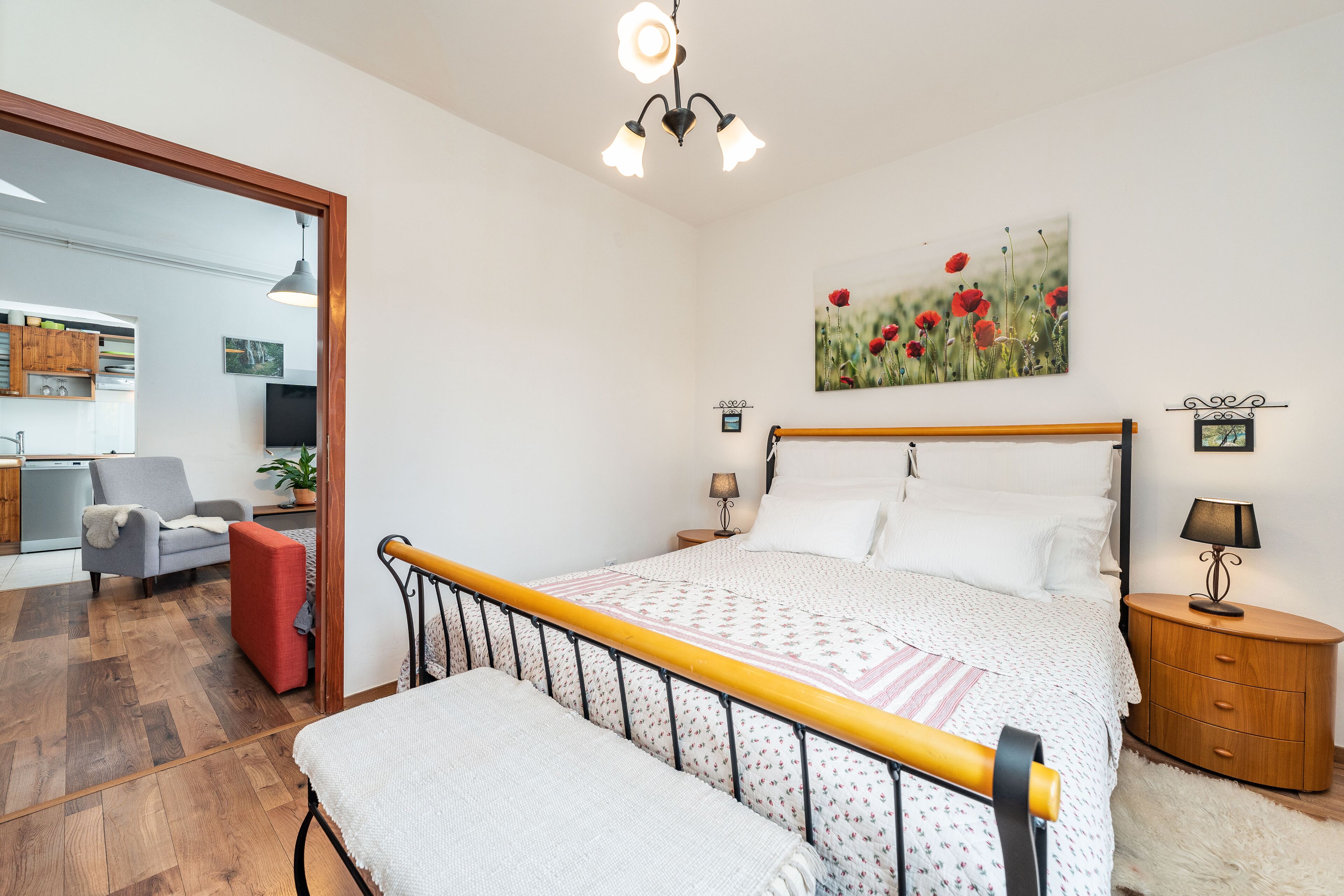 A bedroom with a wooden bed frame, a bedside table with a lamp, a red chair, and a painting of poppies on the wall.