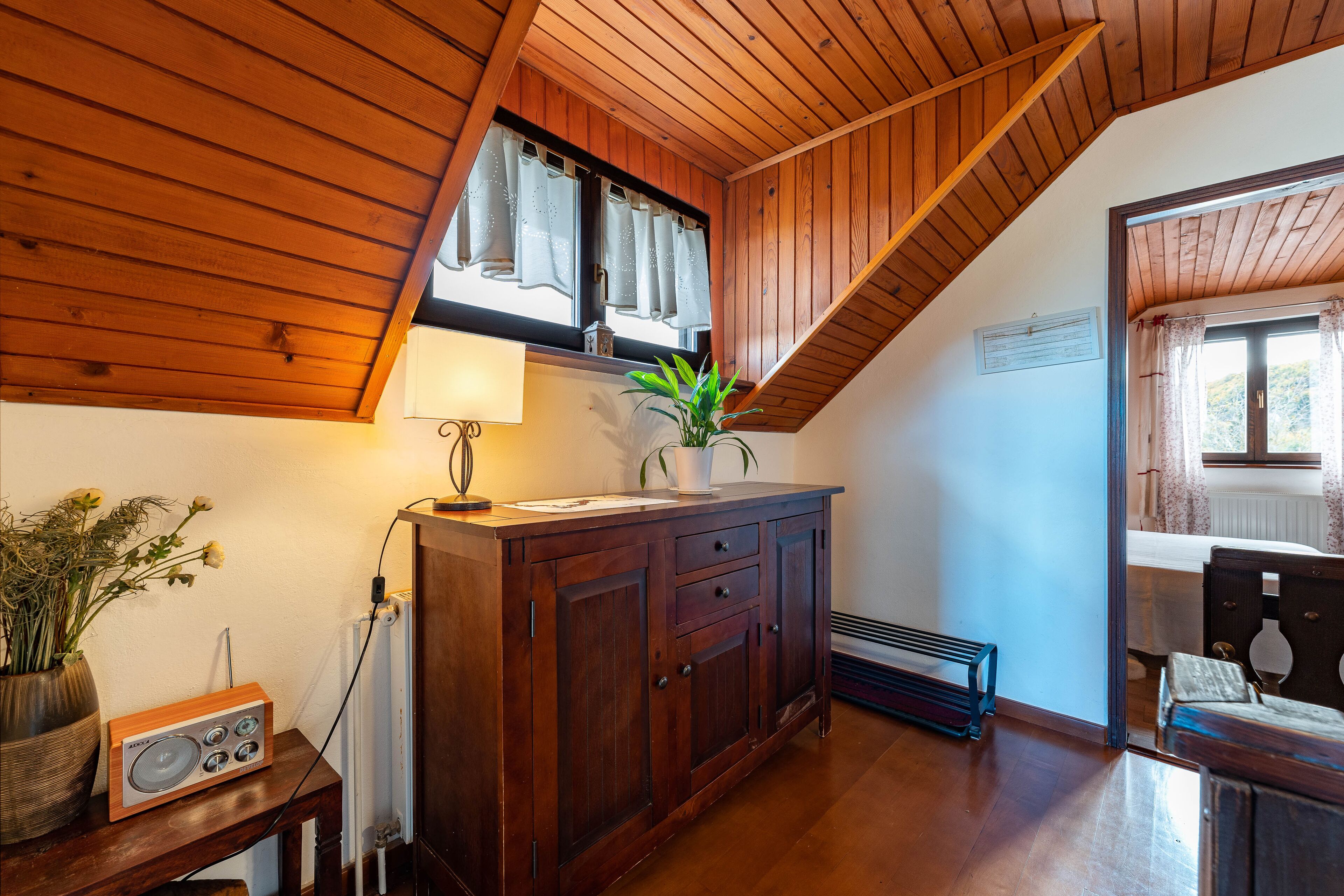Un plafond en bois à pente douce, un meuble en bois avec des tiroirs, un radiateur, une petite table avec une radio et une plante.