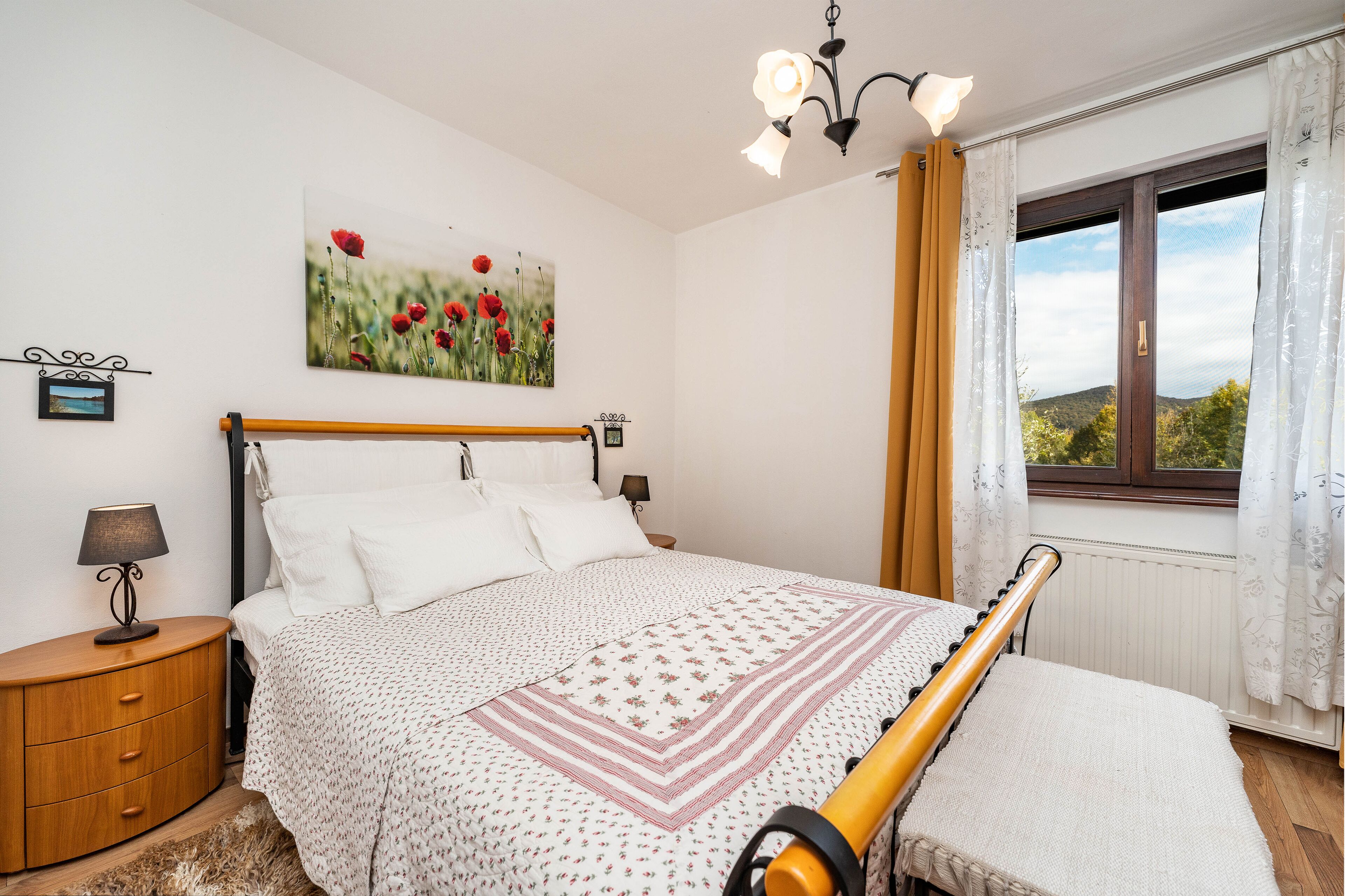 A bedroom with a bed, bedside tables, a lamp, a window with curtains, and a painting on the wall.