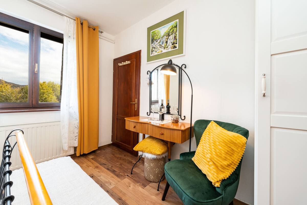 Ein Zimmer mit Holzboden, ein grüner Stuhl mit gelbem Kissen, ein Holztisch mit Spiegel und ein Fenster mit Vorhängen.