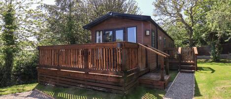A wooden cabin with a deck, surrounded by trees and a gravel path.