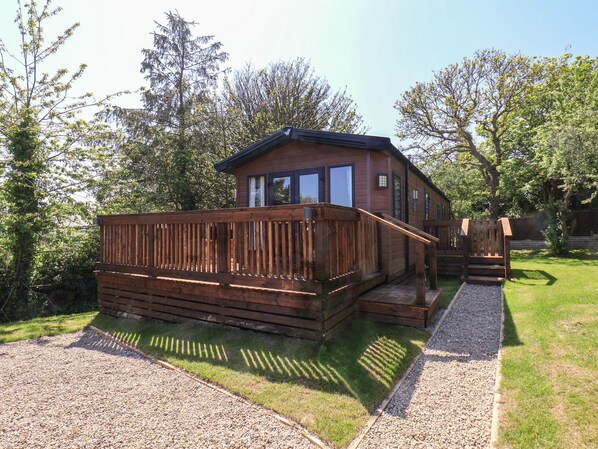 A wooden cabin with a deck, surrounded by trees and a gravel path. - Buttercup Lodge (Saltburn-by-the-Sea)