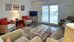 Ein Wohnzimmer mit Korbmöbeln, einem Glastisch, einem Flachbildfernseher und einer großen Schiebetür zum Garten.