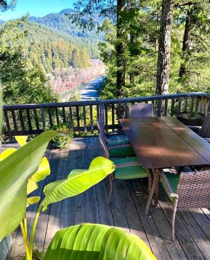 Una terraza de madera con una mesa y sillas, con vistas a un río y montañas boscosas.