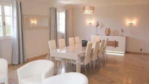 Un vaste espace dédié à la salle à manger, avec une grande table et des chaises blanches, un lustre et des luminaires fixés au mur.