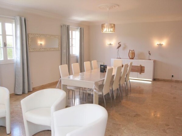 A spacious dining area with a large table and white chairs, a chandelier, and wall-mounted lighting.