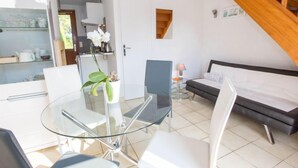 Una habitación moderna con una mesa y sillas de vidrio, una cama, una mesita de noche con lámpara y una escalera.