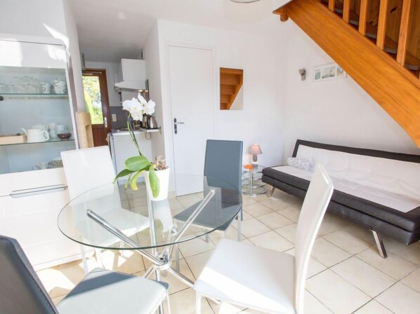 A modern room with a glass table and chairs, a bed, a small table with a lamp, and a staircase.