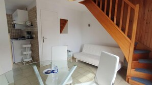 Un espacio habitable compacto con cocina, zona de comedor con mesa de vidrio, sofá blanco y escalera de madera.
