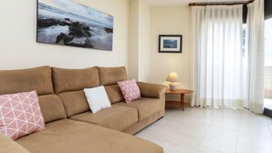 Ein Wohnzimmer mit braunem Sofa, einem kleinen Tisch mit Lampe, einem gerahmten Bild an der Wand und einem großen Fenster mit Vorhängen.