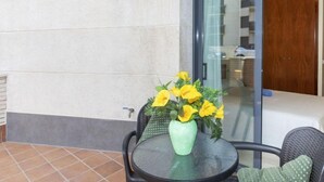 Ein kleiner Außenbereich mit rundem Tisch und Stühlen, einer Vase mit gelben Blumen und einer Glastür zu einem Innenraum.