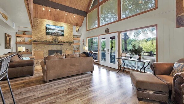 A spacious living room with a fireplace, wooden flooring, and large windows offering a view of the outdoors.