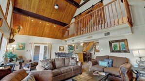 A spacious living room with a high ceiling, wooden beams, a staircase, and a dining area.