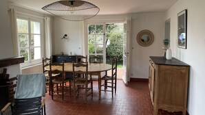 A dining area with a wooden table and chairs, a hanging light fixture, and a view of greenery through open doors. - Le Cottage, Jolie Villa à 3 Minutes de la mer ! (Ambleteuse)