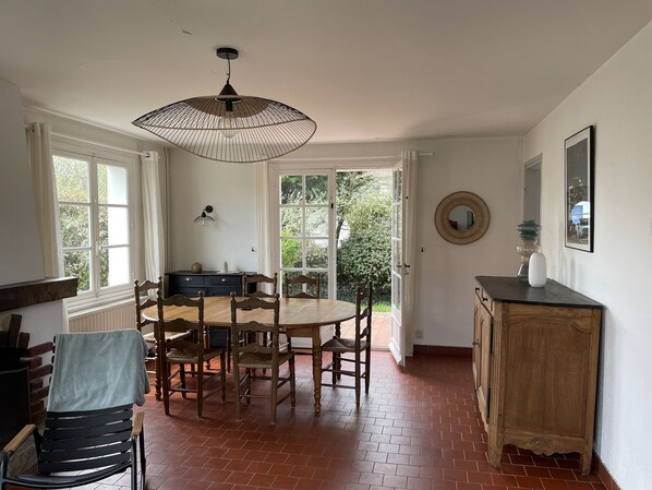 A dining area with a wooden table and chairs, a hanging light fixture, and a view of greenery through open doors.