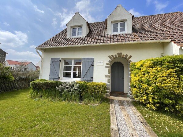 A two-story house with a tiled roof, a front door with an arch, and a sign that reads "LE COTTAGE".