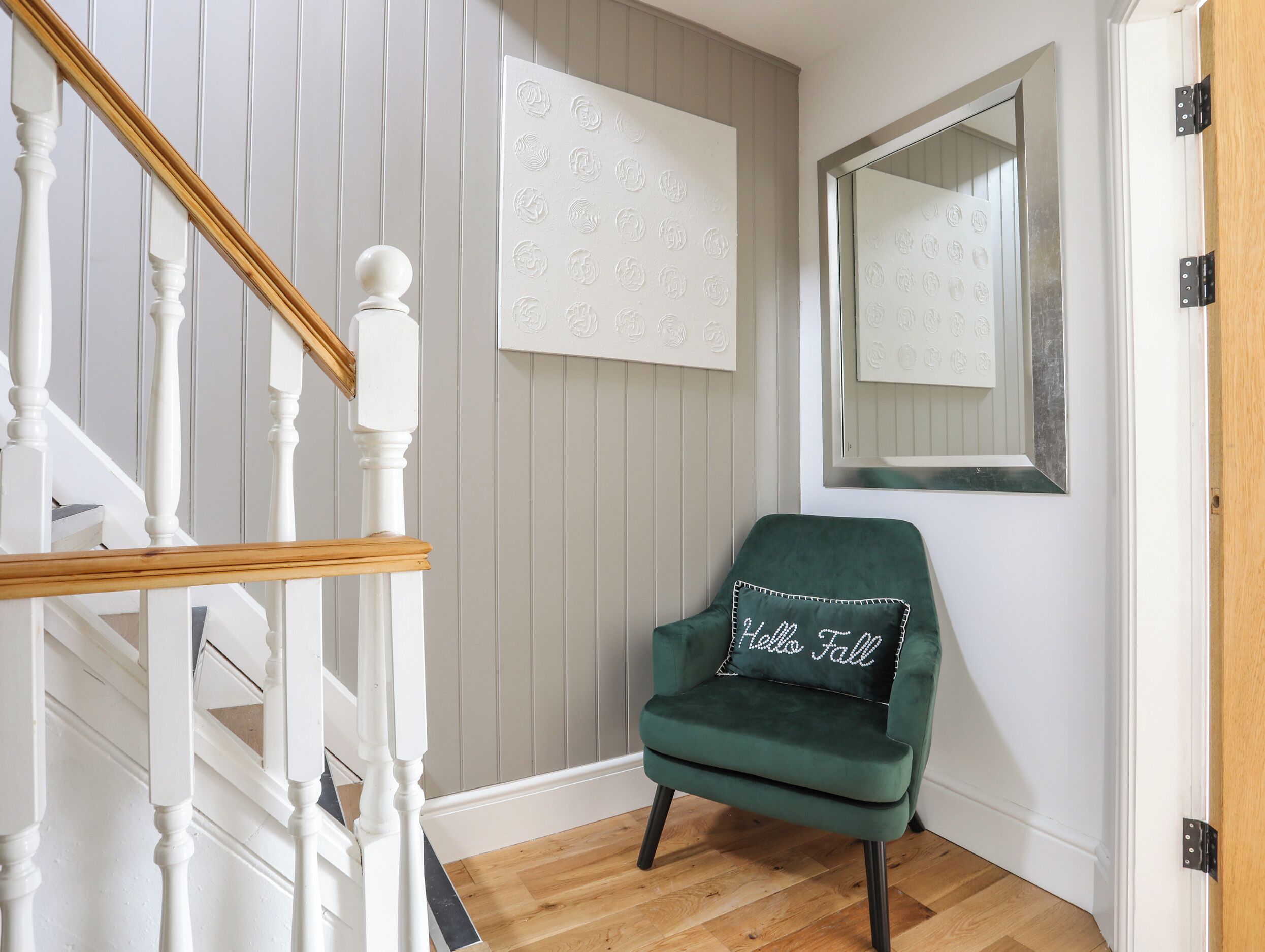 Una escalera con barandillas blancas y un pasamanos de madera, un sillón de terciopelo verde con un cojín que dice 'Hello Fall', una pared blanca con paneles verticales y un espejo con un marco decorativo.