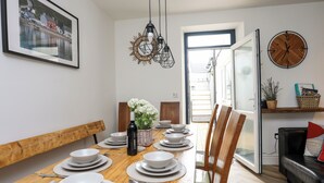 A dining room with a wooden table set for six, white plates, and silverware. There is a framed picture on the wall, a clock, and a window with a view of the outside. - Brigwyn (Pwllheli)