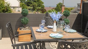 A table set outdoors with a view of the sea, surrounded by greenery and a white building in the background. - Brigwyn (Pwllheli)
