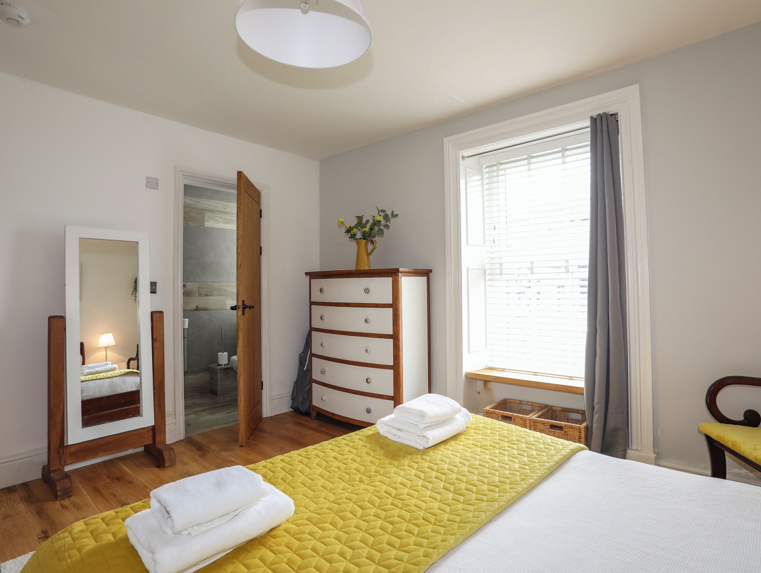 Habitación de hotel con una cama, un aparador de madera, un espejo, una ventana con cortinas y una puerta que da a un baño.