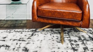 Una habitación con un sillón de cuero marrón, una mesa de centro de vidrio y una lámpara negra, junto con dos cuadros abstractos en la pared.