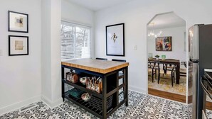 Un coin repas avec une table et des chaises en bois, un meuble en métal noir contenant des ustensiles de cuisine, et un sol à motifs.