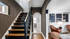 Una escalera moderna con escalones de madera y barandilla negra, que lleva a un pasillo con un tapete estampado y un otoman redondo.