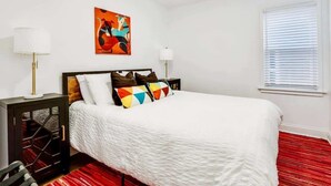 Une chambre à coucher avec un lit, deux tables de chevet, une fenêtre avec des stores et une peinture abstraite colorée accrochée au mur.