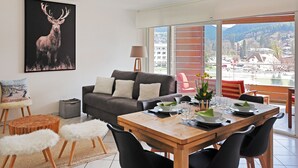 A modern living room with a large window, a dining area with a wooden table, and a sofa.