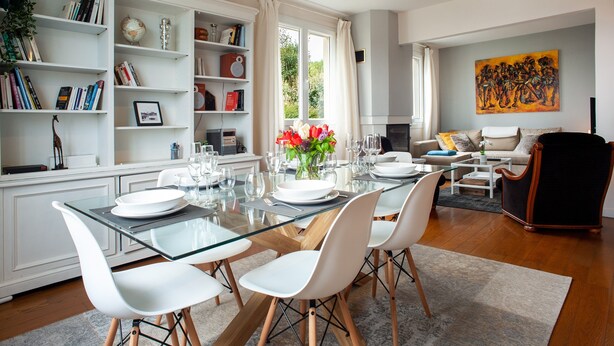 A modern living room with a glass dining table, white chairs, a bookshelf, and a painting on the wall. - Le Gîte de Beauport - Proche des plages (Paimpol)