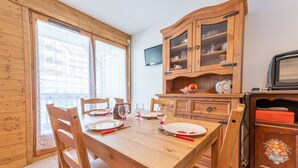 A wooden dining table set with plates, glasses, and cutlery. A wooden cabinet with glass doors is against the wall, and a television is mounted above it.