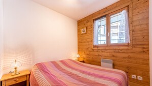 A bedroom with a wooden wall, a window with a curtain, a bed with a striped bedspread, a nightstand with a lamp, and a picture on the wall.