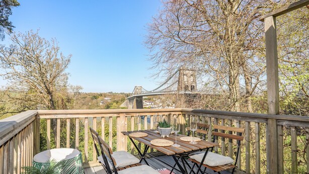 Une terrasse en bois avec une table et des chaises, d’où l’on a vue sur un pont et des bâtiments.