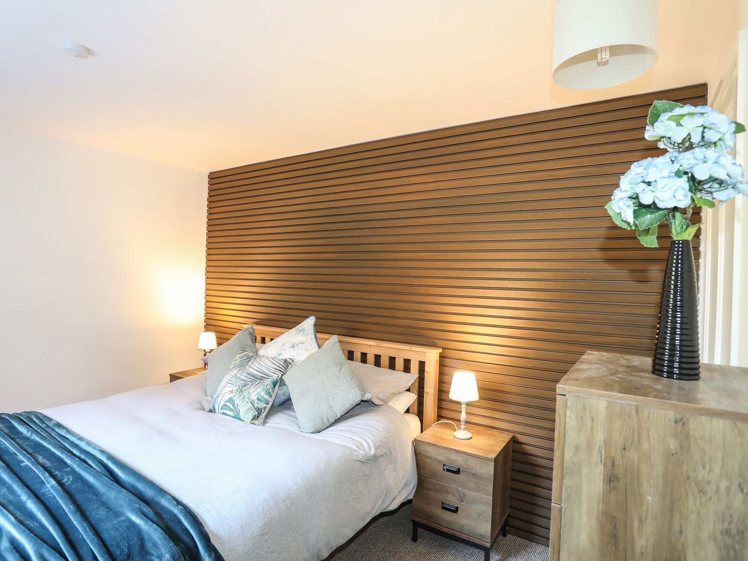 A bedroom with a bed, bedside tables, a nightstand, a lamp, and a vase with flowers.
