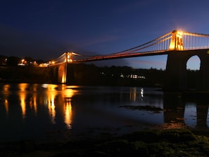 Vista noturna de uma ponte sobre um rio calmo, com suas luzes refletindo na água.