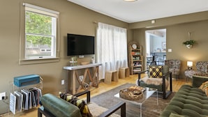 A living room with a television, a table, and a sofa. There is a window with a view of trees and a ceiling light.